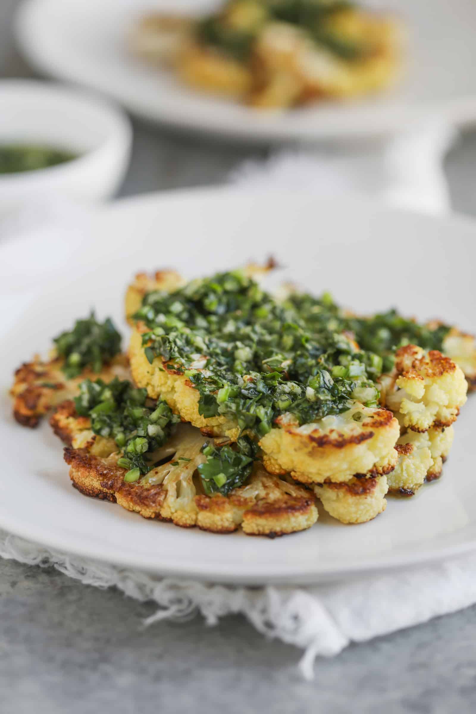Roasted Cauliflower Steaks with Chimichurri Sauce 10 Roasted Cauliflower Steaks Topped With Vibrant Chimichurri Sauce Are Served On A White Plate. The Dish Is Elegantly Presented On A Grey Surface, Accompanied By A Bowl Of Extra Sauce In The Background.