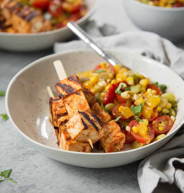 Grilled Chicken Skewers Sit In A Bowl With A Colorful Corn, Tomato, And Avocado Salad, Perfect For A Healthy Cookout. The Dish Is Garnished With Fresh Herbs. A Fork Rests In The Bowl, And A Cloth Napkin Is Nearby. In The Background, Another Bowl Of Vibrant Salad Completes The Scene.