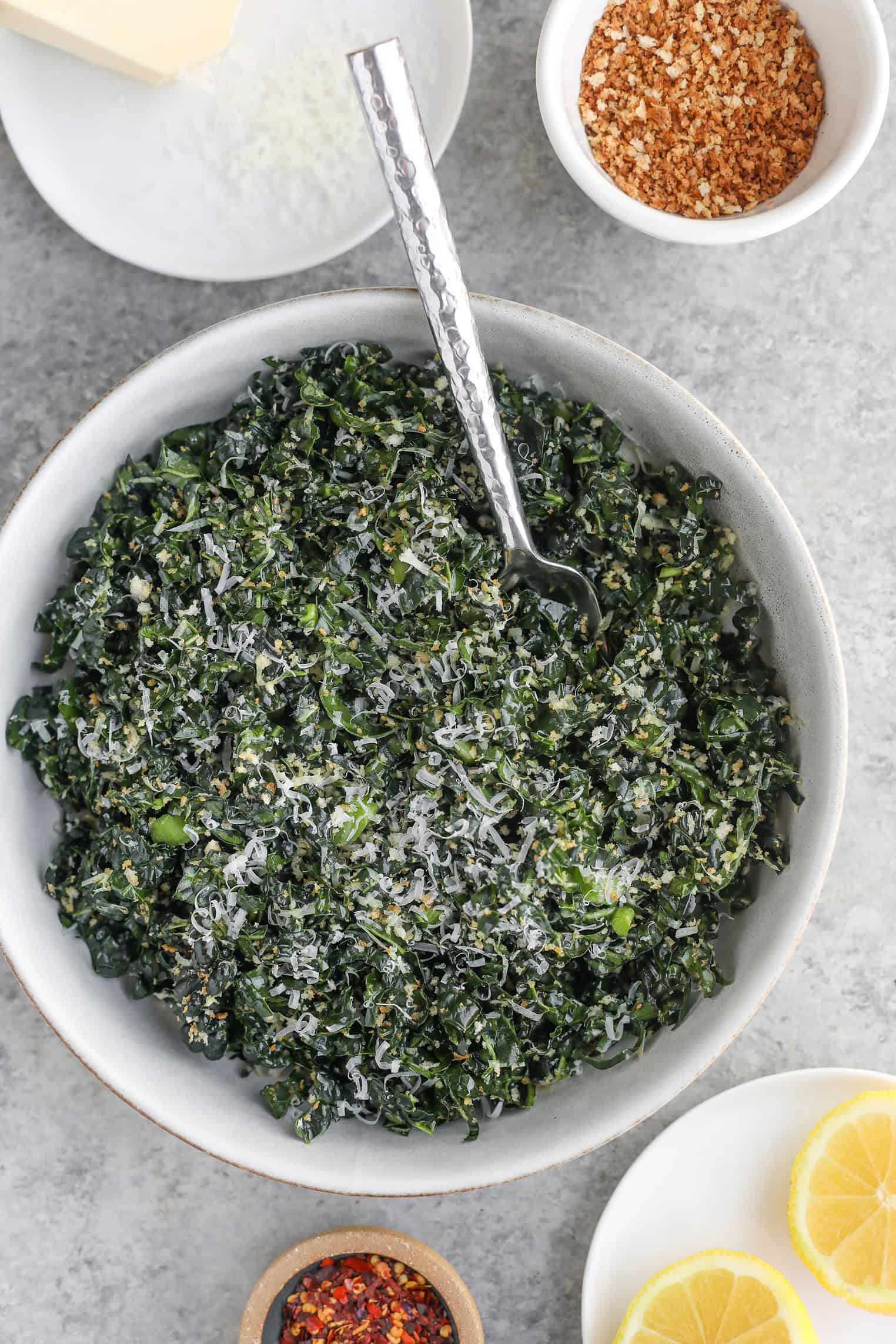 Easy Kale Caesar Salad (6 Ingredients!) 8 A Bowl Of Kale Caesar Salad Topped With Grated Cheese And A Metallic Spoon Sits Nearby. Small Bowls Hold Breadcrumbs, Red Chili Flakes, And Butter. Two Lemon Slices Are Arranged On A White Plate Beside Them, All Displayed On A Gray Surface.