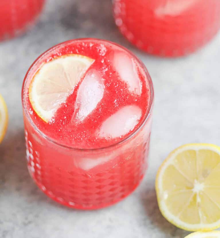 A Glass Filled With A Bright Pink Drink And Ice Cubes, Garnished With A Lemon Slice, Sits On A Gray Surface. Two Lemon Halves Are Nearby, Enhancing The Fresh And Summery Vibe Of This Healthy Vegan Cookout Scene.