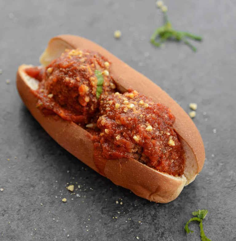 A Hoagie Roll Filled With Two Meatballs, Topped With Marinara Sauce And Sprinkled With Grated Cheese, Sits On A Dark Surface. Scattered Herbs Are Visible Around The Sandwich, Reminiscent Of Flavors From Healthy Vegan Cookout Recipes.