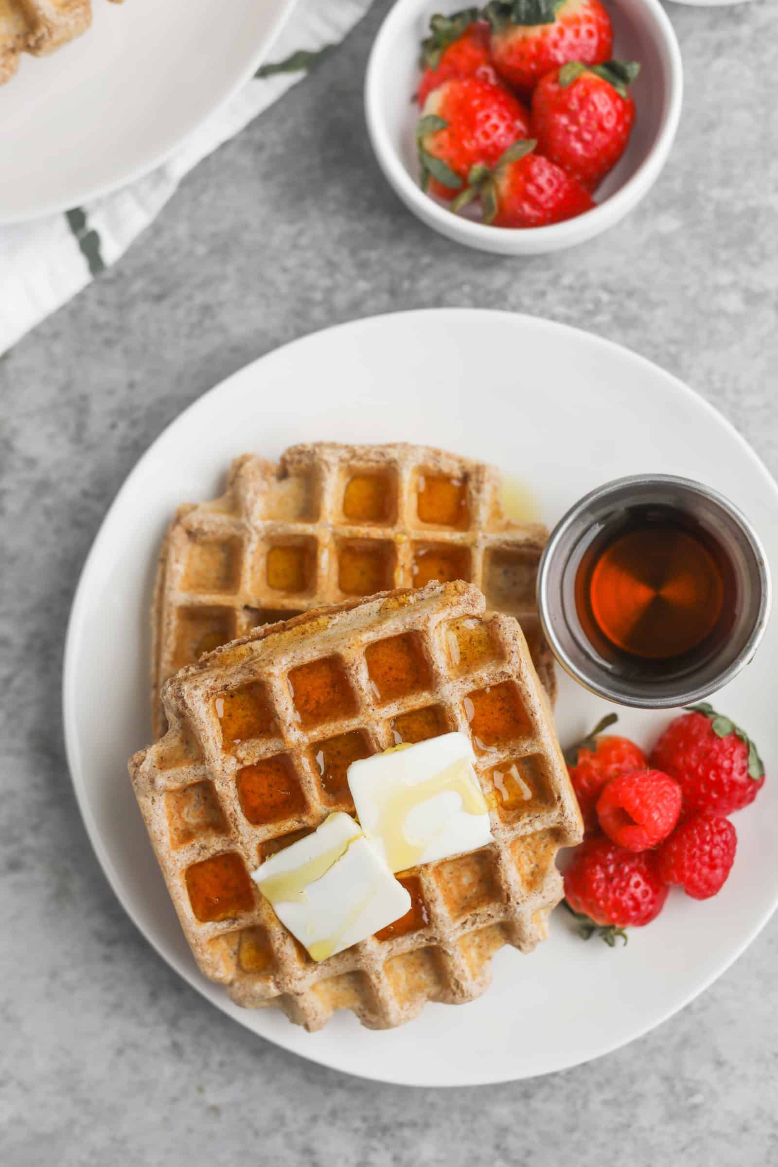 Whole Wheat Waffles 13 Two Whole Wheat Waffles With Butter And Syrup Rest On A White Plate, Accompanied By A Small Dish Of Syrup And A Few Strawberries. Nearby, Another Bowl Brims With Strawberries. The Backdrop Is A Textured Gray Surface, Creating A Cozy Breakfast Setting.