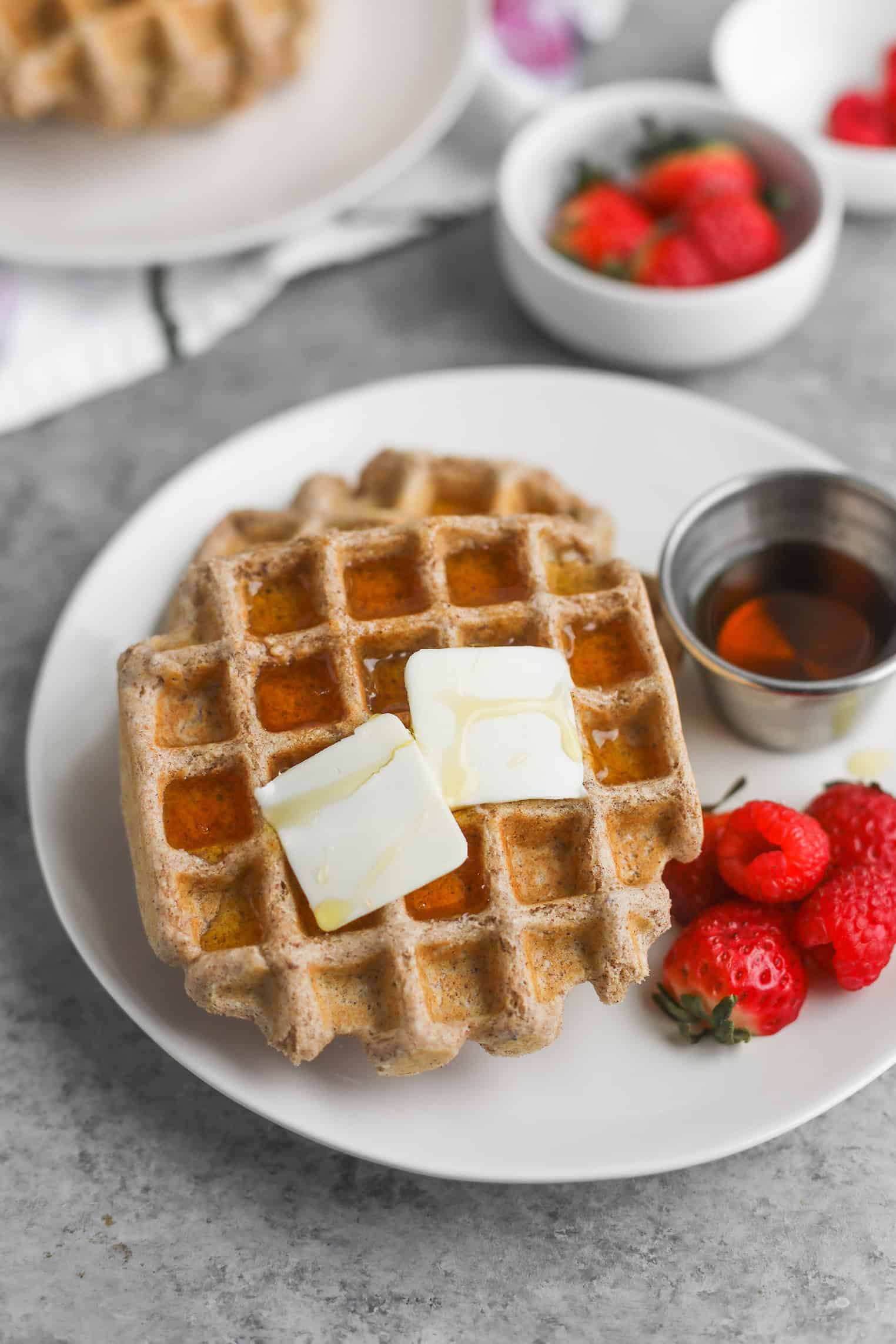 Whole Wheat Waffles 11 A Plate With Two Whole Wheat Waffles Topped With Melting Butter And Drizzled With Syrup. A Small Cup Of Syrup And Fresh Strawberries And Raspberries Sit On The Side, All Set Against A Gray Surface.