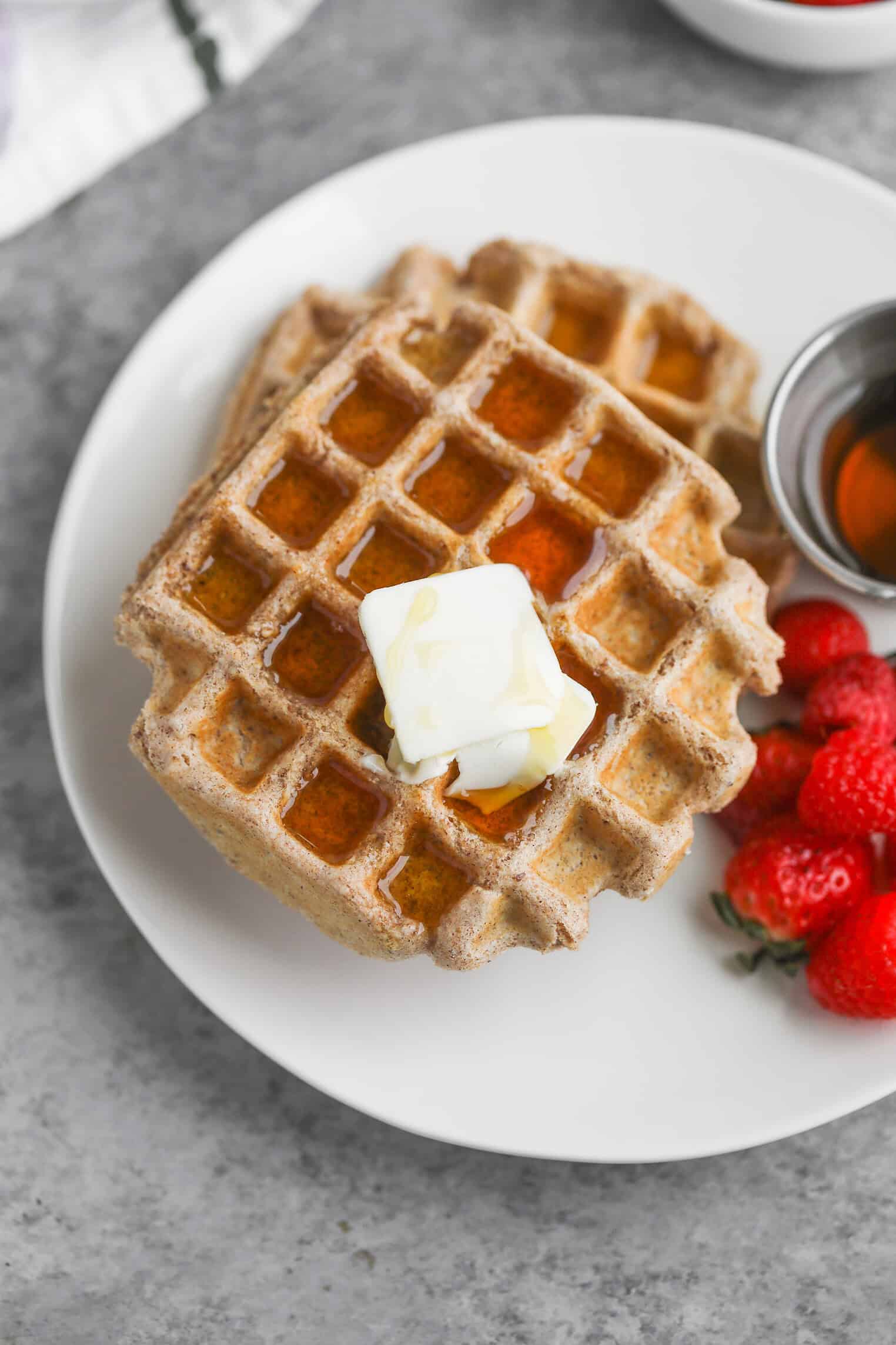 Whole Wheat Waffles 2 Whole Wheat Waffles Topped With A Pat Of Butter And Syrup On A White Plate, Accompanied By Fresh Strawberries And A Small Metal Cup Of Syrup, Set On A Gray Surface.