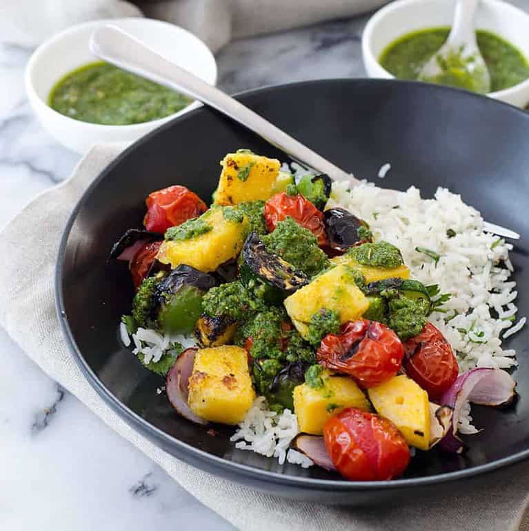 A Black Bowl Contains White Rice Topped With Grilled Vegetables Like Cherry Tomatoes, Zucchini, And Red Onions. It'S Garnished With Green Pesto Sauce, Perfect For Healthy Vegan Cookout Recipes. A Fork Rests On The Bowl. In The Background, Two Small White Bowls Hold Extra Green Sauce.
